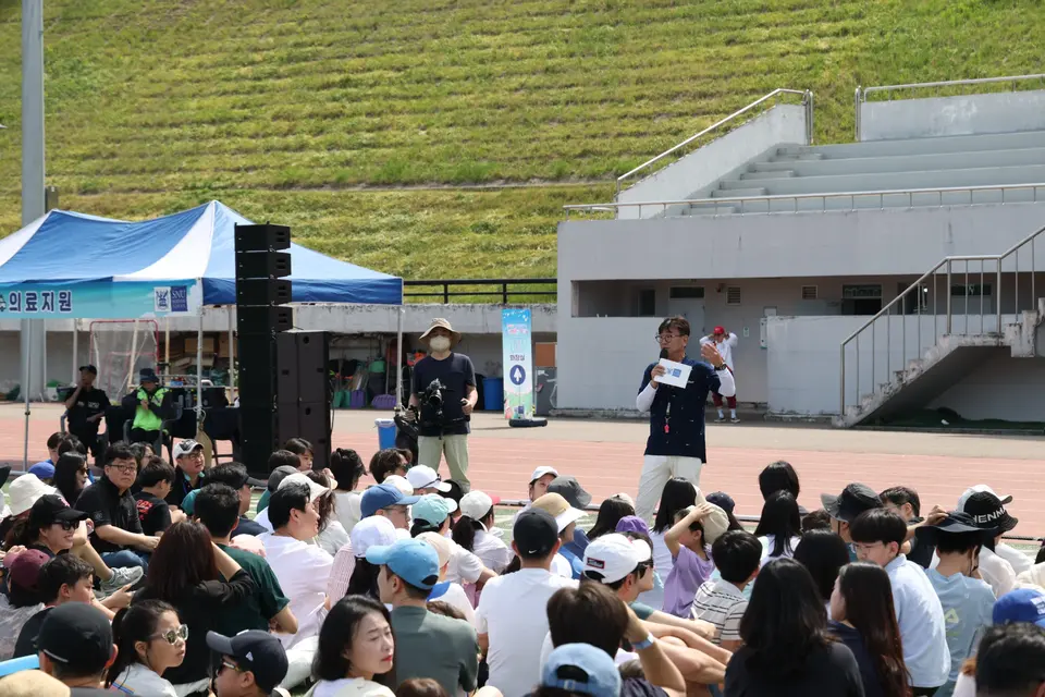운동장 스탠드 앞 무대에서 진행하는 MC와 앉아 있는 참가자들