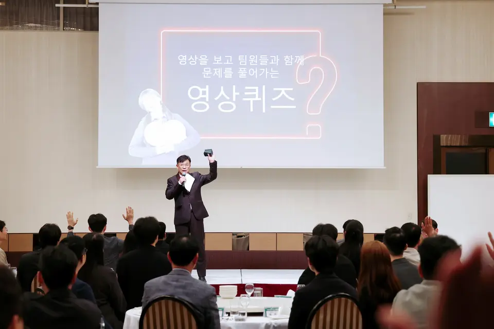 영상 퀴즈 화면 앞에서 손을 들어 진행하는 MC와 연회장 객석